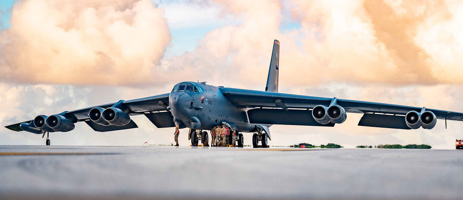 A large military aircraft with wide wings stretches across a runway, set against a backdrop of colorful clouds. Crew members gather at the nose, conveying teamwork.