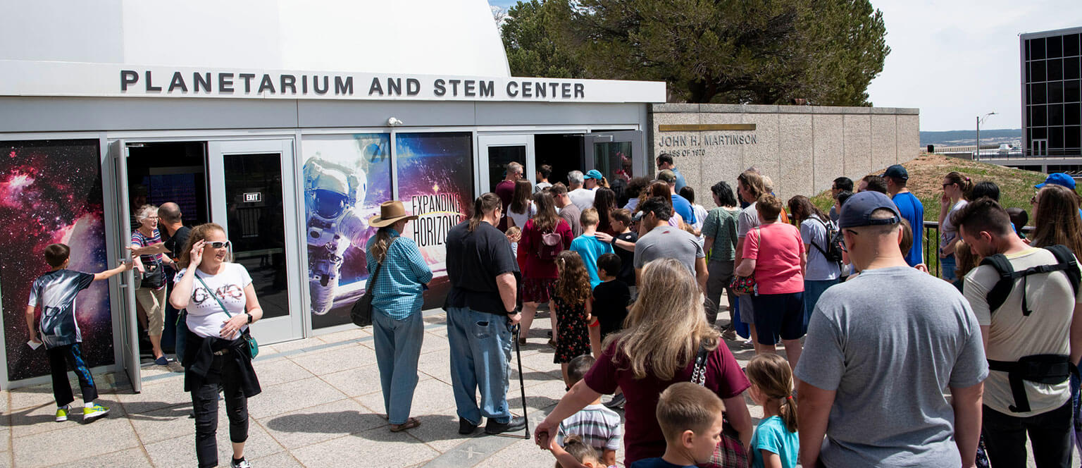 A large group of people, including adults and children, waits in line outside the Air Force Academy's Planetarium and STEM Center.