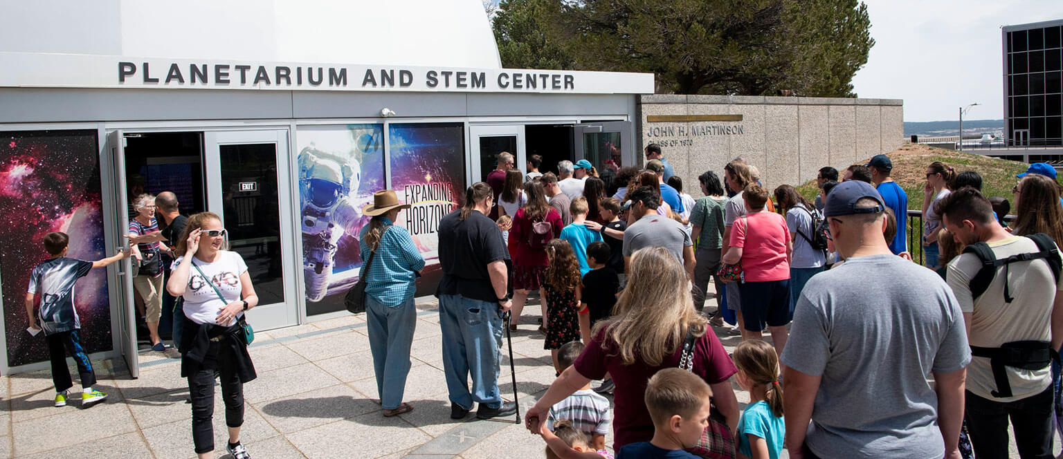 A crowd, including families, waits outside the U.S. Air Force Academy planetarium and STEM center under a clear sky. The atmosphere is lively and anticipatory.