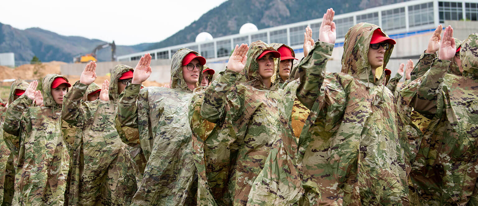 A group of basic cadets stands outdoors, raising their right hands during swearing-in. They wear camouflage ponchos and red caps, conveying discipline and unity against a mountainous backdrop.