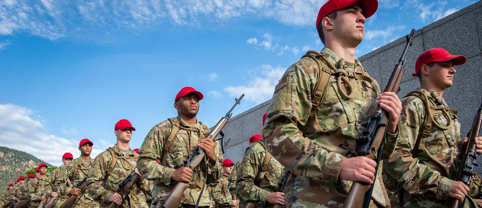 A group of basic cadets in camouflage uniforms and red caps march in formation under a blue sky. Each carries a rifle, conveying discipline and unity.