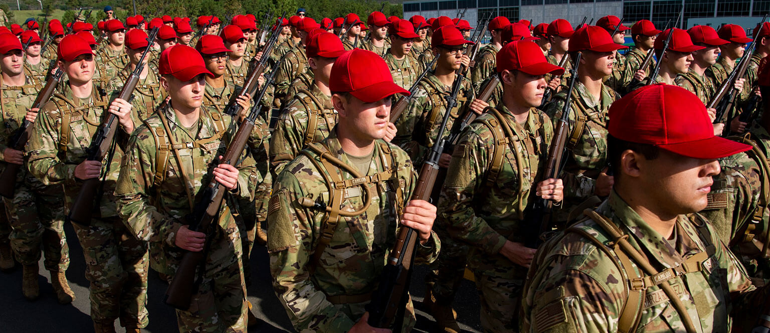 A group of basic cadets in camouflage uniforms and red caps march in unison, holding rifles vertically. The image conveys discipline and unity.