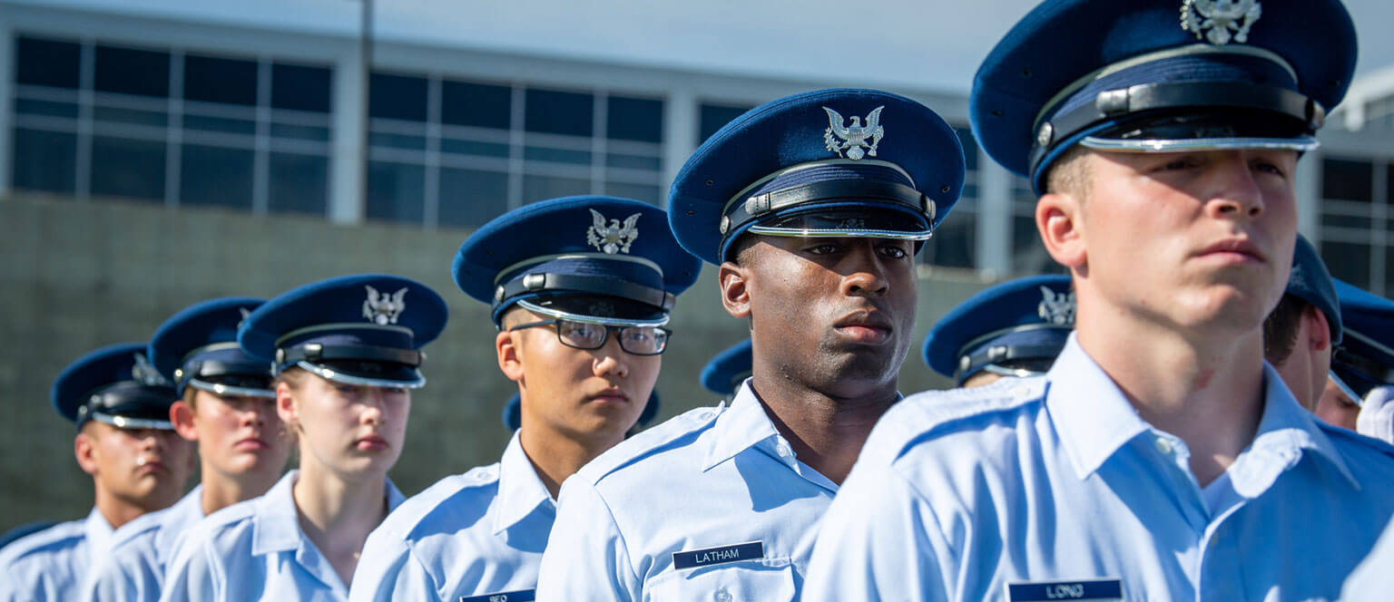 Cadets basics in uniform and blue caps stand in crisp rows during a ceremony, appearing focused and disciplined, with a building in the background.