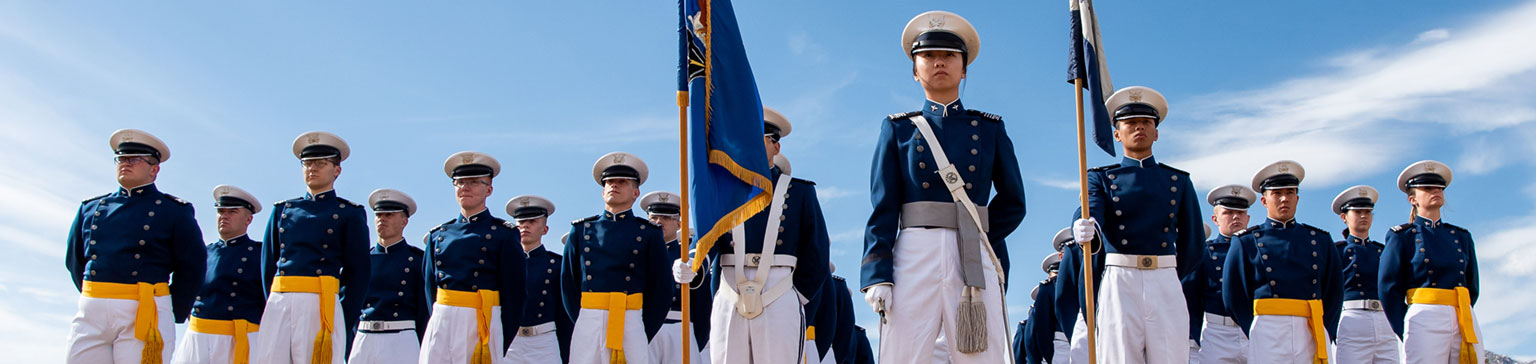 A group of cadets in parade uniforms stand in formation, holding flags against a clear blue sky, exuding discipline and unity.
