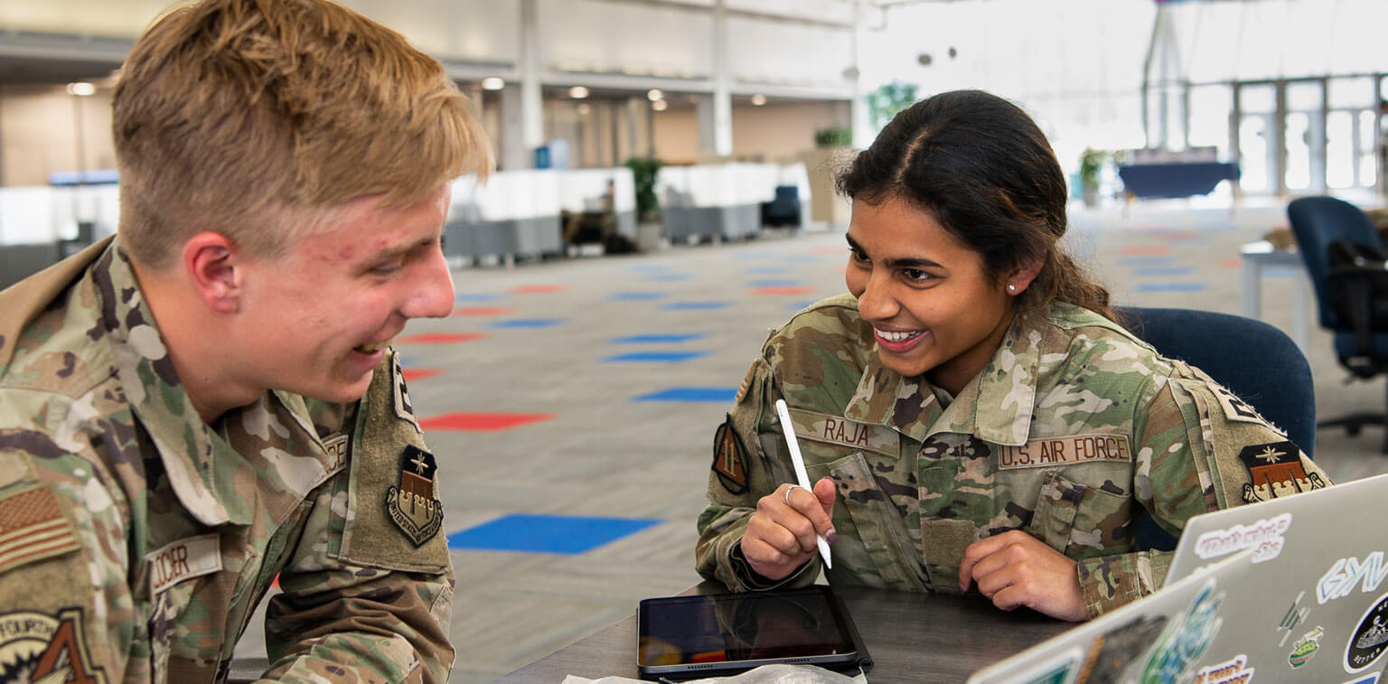 Two cadets personnel in camouflage uniforms smile and converse in an open office space. They sit at a table with a tablet and a laptop nearby.