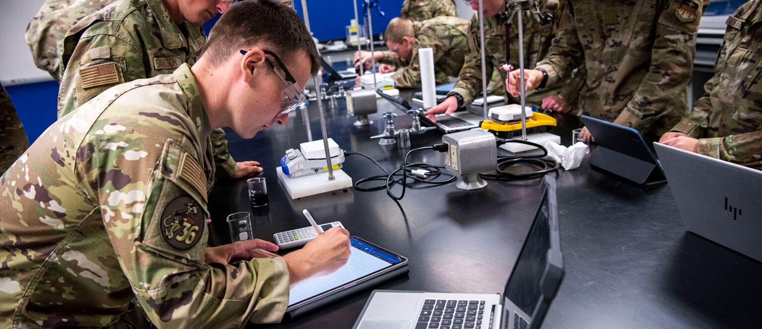 Cadets in camouflage uniforms conduct experiments in a lab, focusing on scientific equipment and using tablets. The atmosphere is serious and attentive.