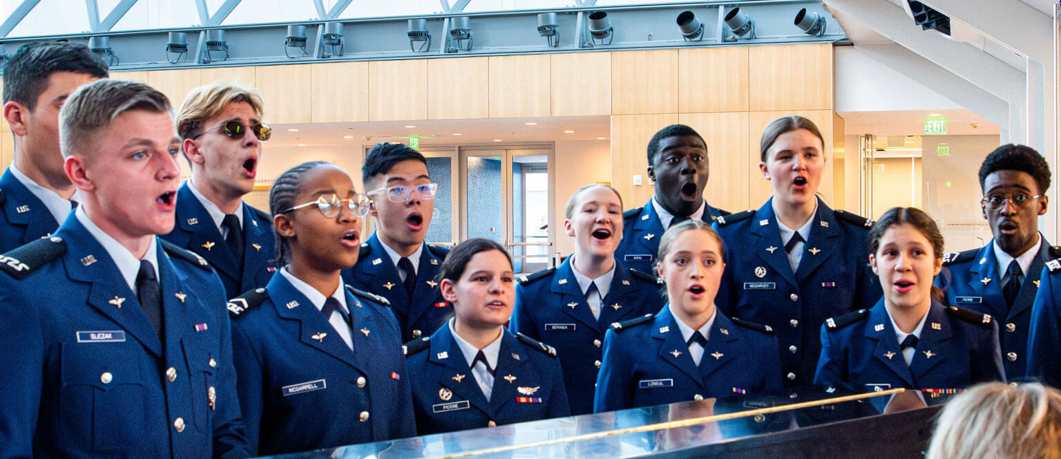 A group of U.S. Air Force Academy cadets singing in the cadet chorale enthusiastically in a Polaris Hall. They show focus and passion, standing close together in a bright, airy space.