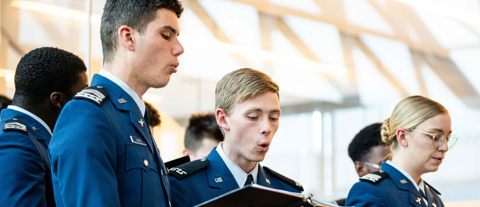 A group of uniformed cadets sing from songbooks in a bright, airy space. The mood is focused and composed as they participate in a formal event.