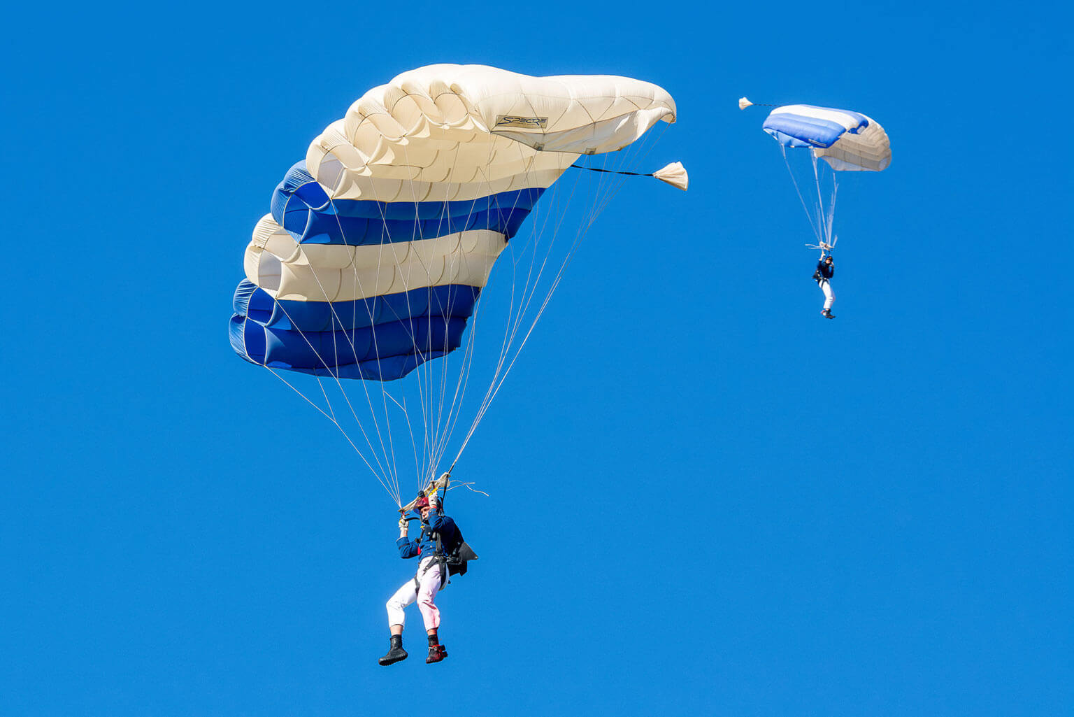 Two Wings of Blue members with blue and white parachutes gracefully descend into Stillman Field against a clear blue sky. Two Wings of Blue members with blue and white parachutes gracefully descend into Stillman Field against a clear blue sky.