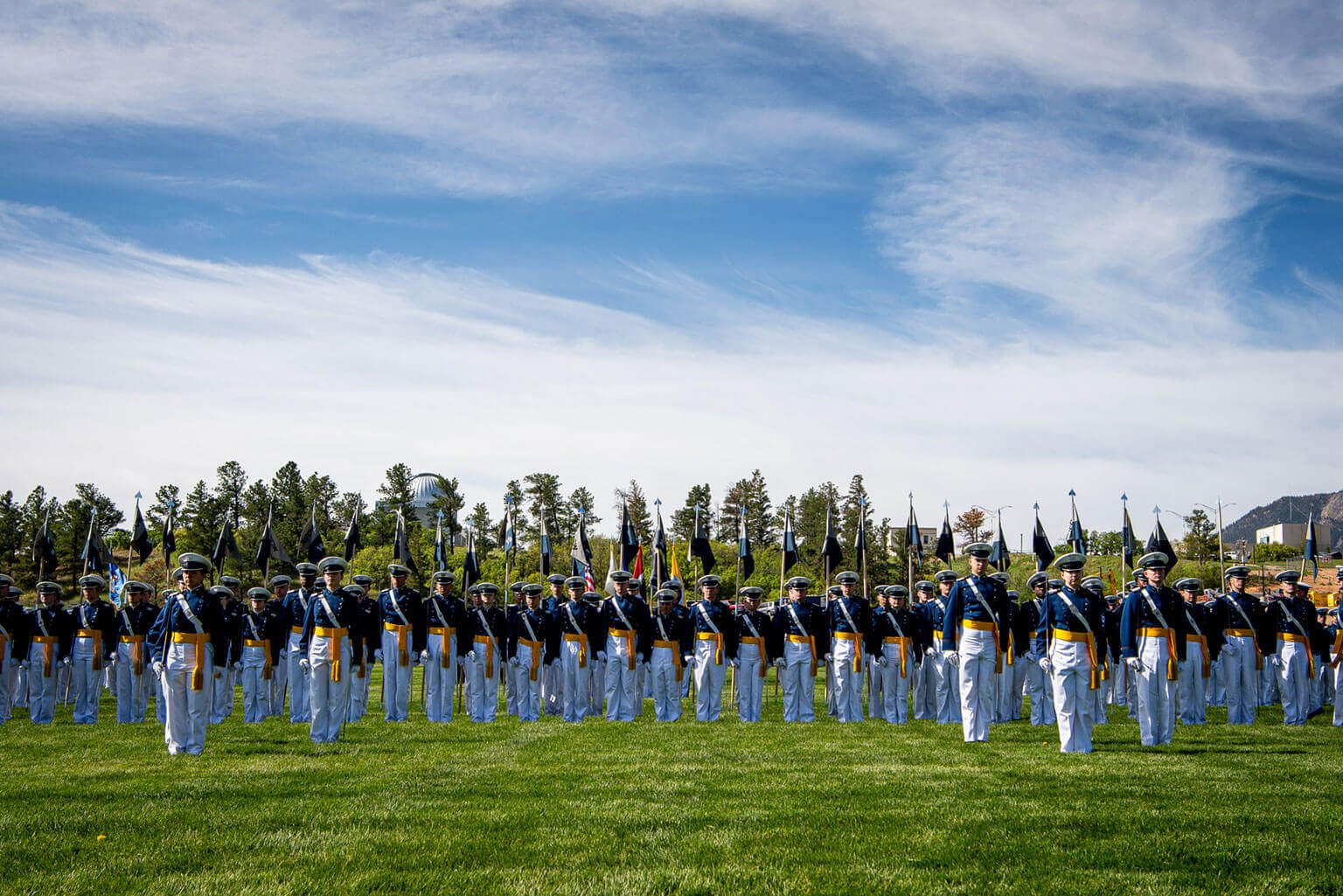 A large group of cadets in dress uniforms stand in formation on Stillman Field under a blue sky with scattered clouds. The scene is in a spacious, outdoor area with trees in the background. A large group of cadets in dress uniforms stand in formation on Stillman Field under a blue sky with scattered clouds. The scene is in a spacious, outdoor area with trees in the background.