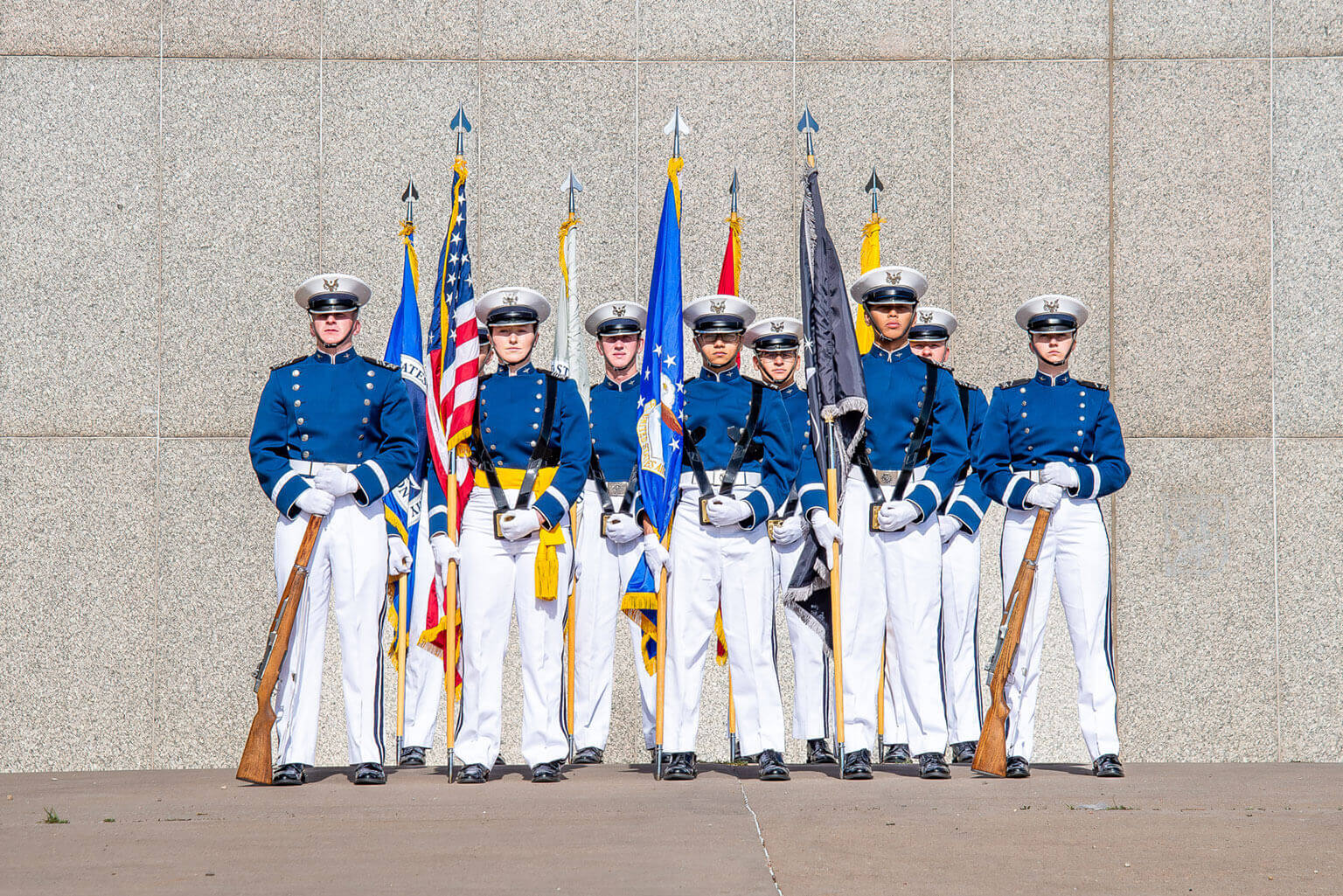 A group of uniformed cadets stands in formation against a concrete wall, poised for the Graduation Parade. They hold rifles and flags, including the American flag, ready to present the colors. The cadets wear blue jackets, white pants, and caps as the sun casts shadows on the ground. A group of uniformed cadets stands in formation against a concrete wall, poised for the Graduation Parade. They hold rifles and flags, including the American flag, ready to present the colors. The cadets wear blue jackets, white pants, and caps as the sun casts shadows on the ground.
