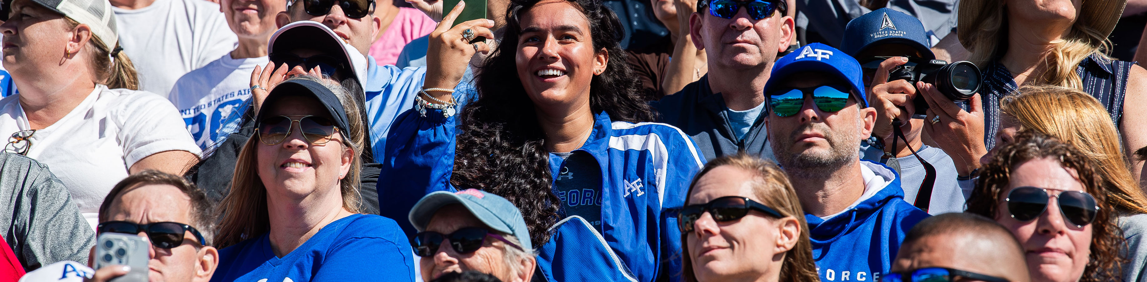 A diverse crowd of people, mostly in blue apparel, are sitting outdoors, smiling and cheering enthusiastically, creating a lively and energetic atmosphere.