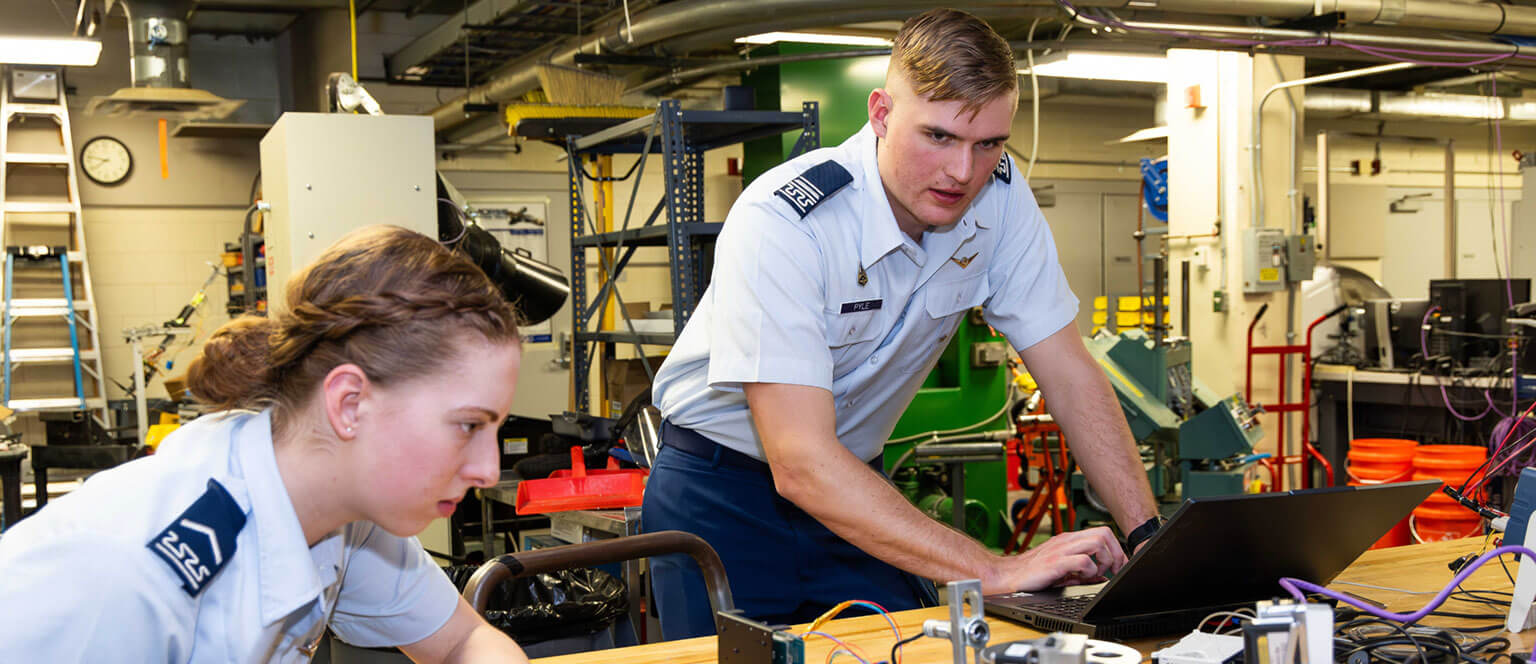 Two cadets work on a technical project in a lab. One operates a laptop, while the other focuses on equipment. The setting is industrious and collaborative.