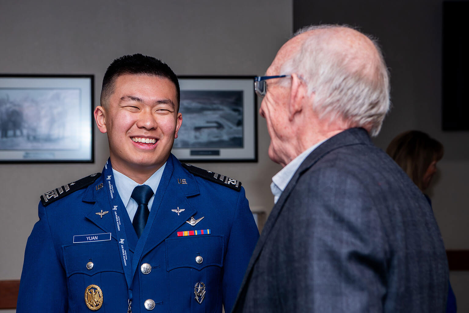 A U.S. Air Force Academy cadet smiles while talking to an older man in a suit jacket at an indoor event. Framed photos hang on the wall in the background.