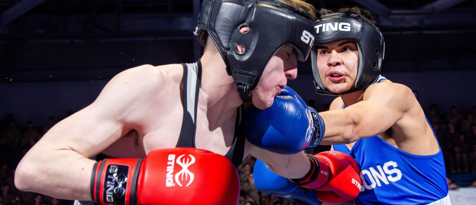 Two cadets in protective headgear are mid-fight; one in blue lands a punch on the other's face while both wear boxing gloves and athletic attire.