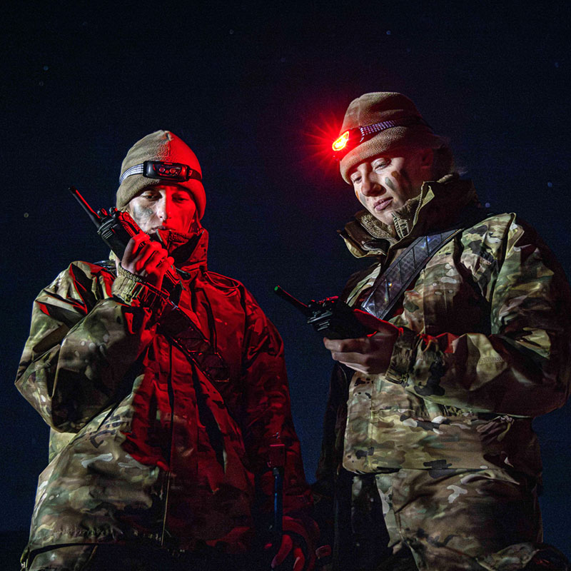 Two cadets in camouflage gear and headlamps stand at night, communicating with handheld radios under red light.