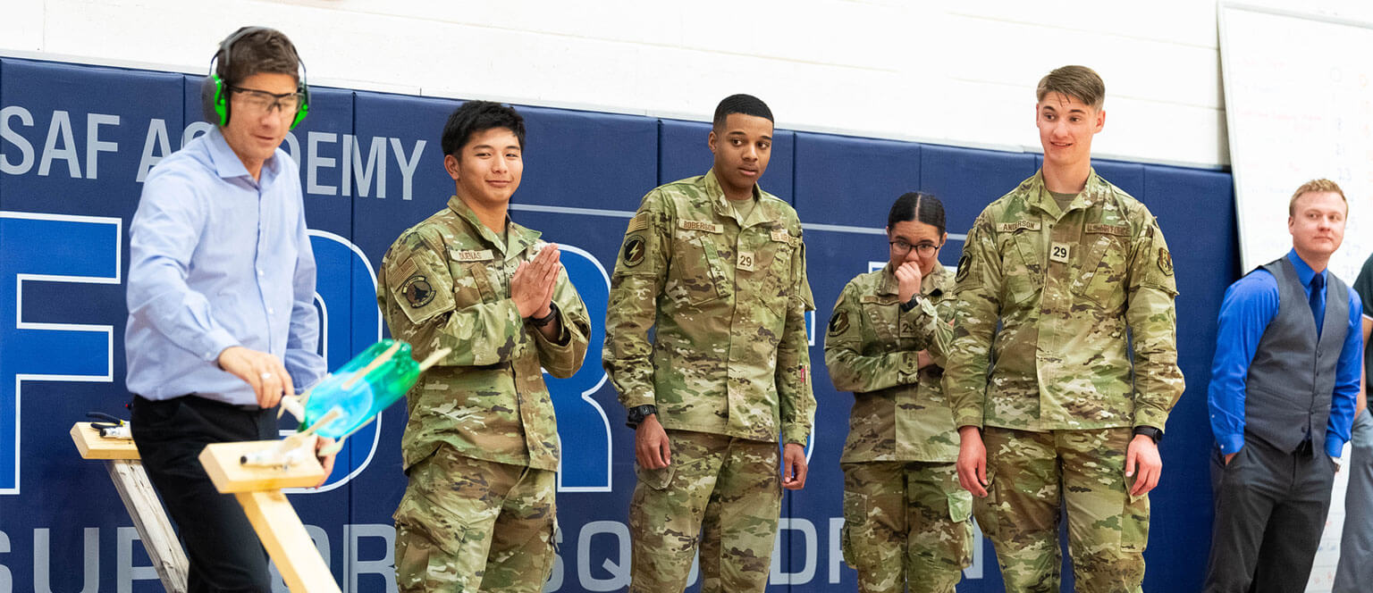 A group of prep school cadet candidates stand in front of a wall, observing an experiment being conducted.