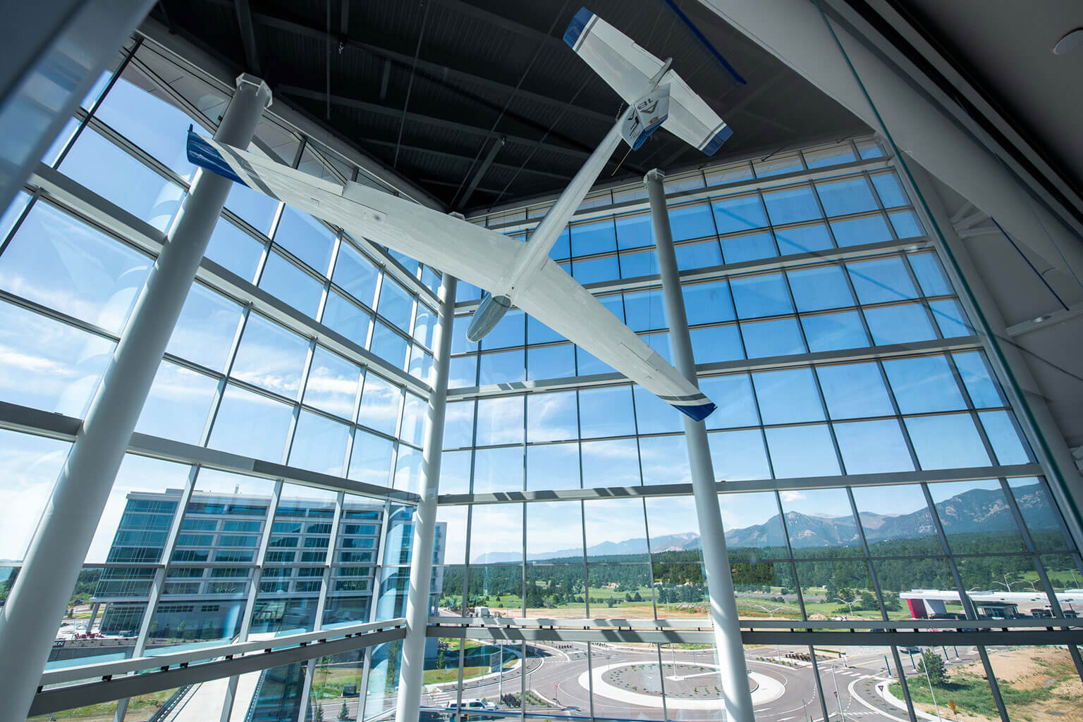 A suspended saillane is displayed within a large, modern glass atrium. The sunny view outside shows green fields, buildings, and distant mountains, creating an inspiring and expansive feel.