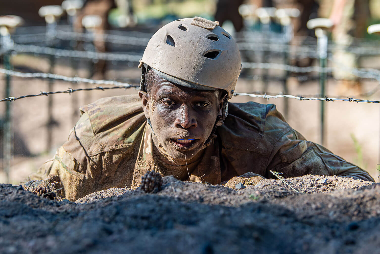 A basic cadet in camo gear and helmet crawls under barbed wire in a military training exercise. The expression is focused, with determination evident. 