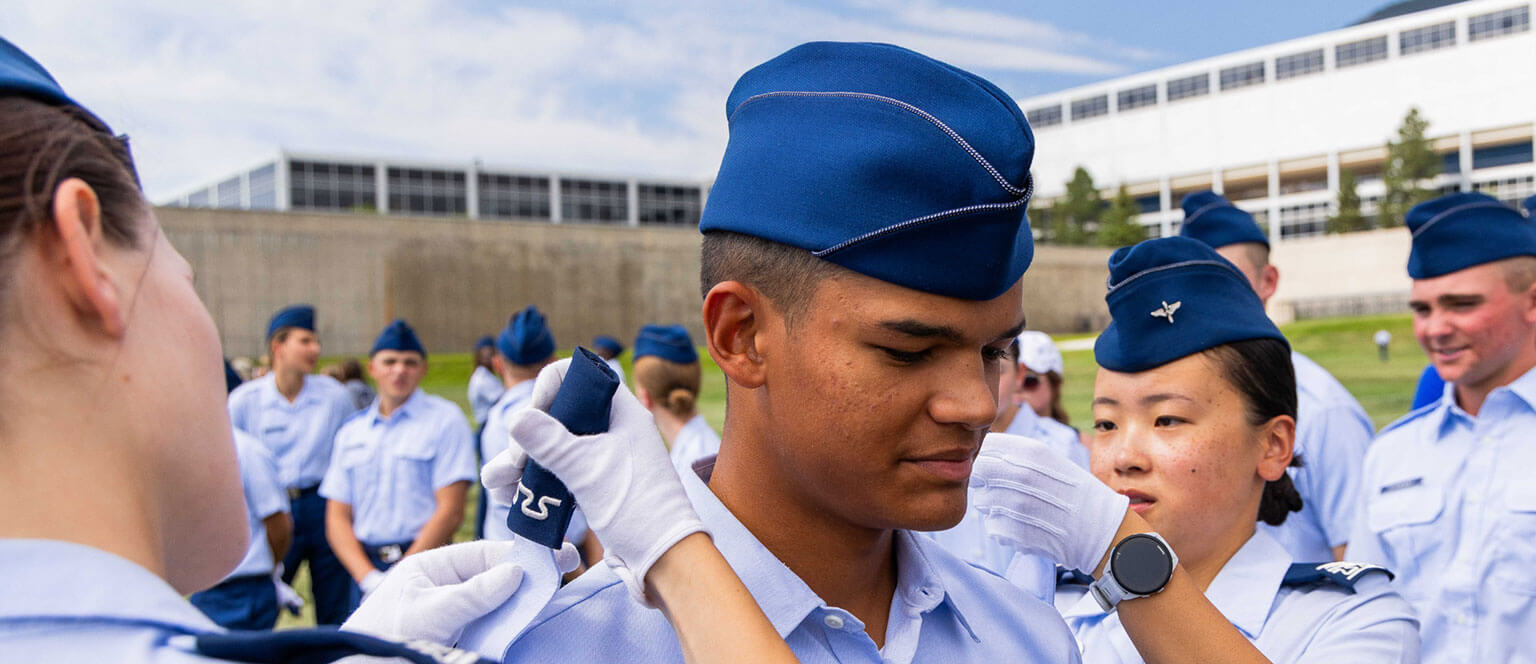 Air Force Cadets in blue uniforms participate in an outdoor ceremony. One cadet receives shoulder boards while others look on. A building is visible in the background under a blue sky.