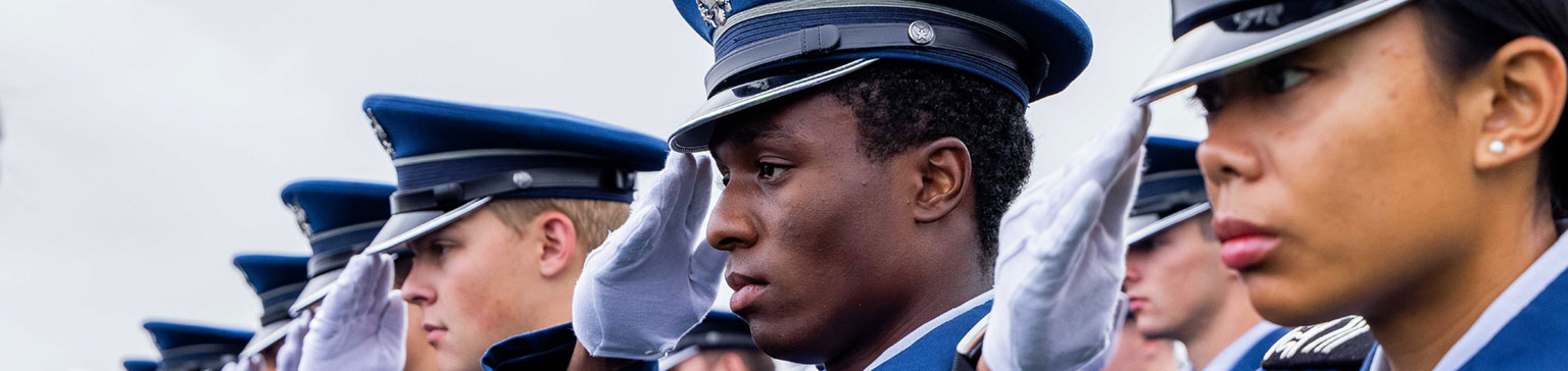 A group of cadets in uniform stands in formation, saluting. Their expressions are focused and serious, conveying discipline and respect.