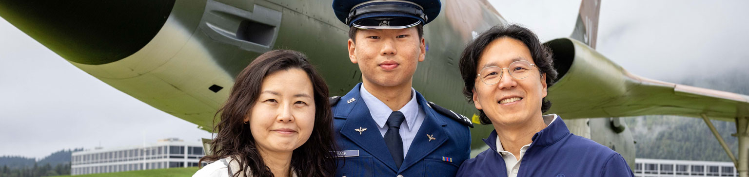 A male cadet in a blue uniform stands proudly with his parents in front of an F-105D Thunderchief on a cloudy day, evoking pride and joy.