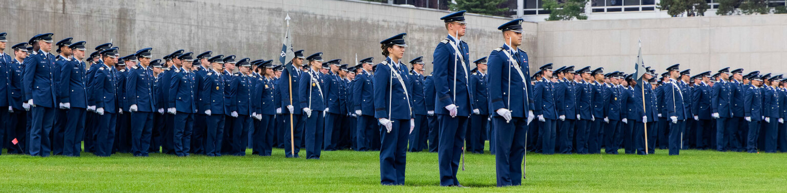 Cadets formed by squadron stand at attention on Stillman Field. They wear blue uniforms and hats, conveying discipline and formality.