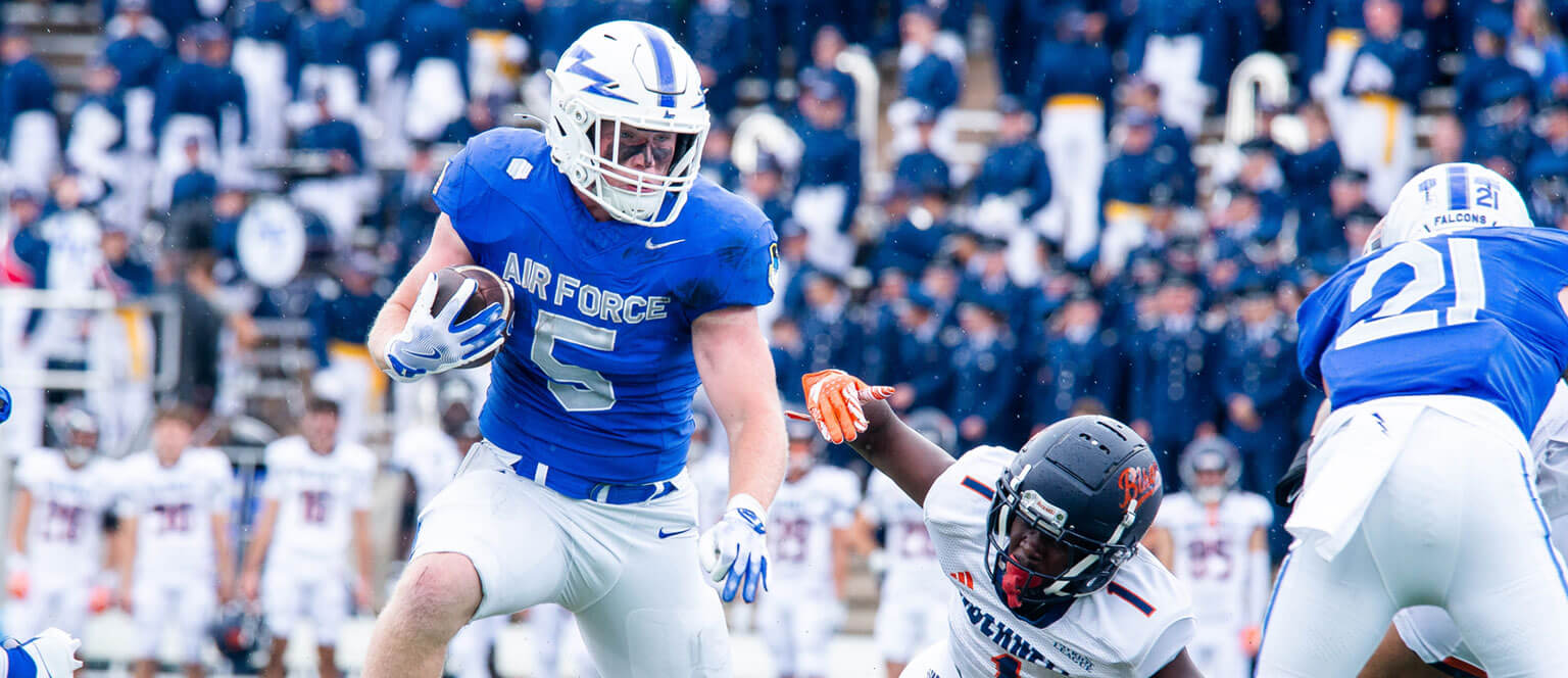A U.S. Air Force Academy football player in motion, carrying the ball as he navigates past defenders on the field.