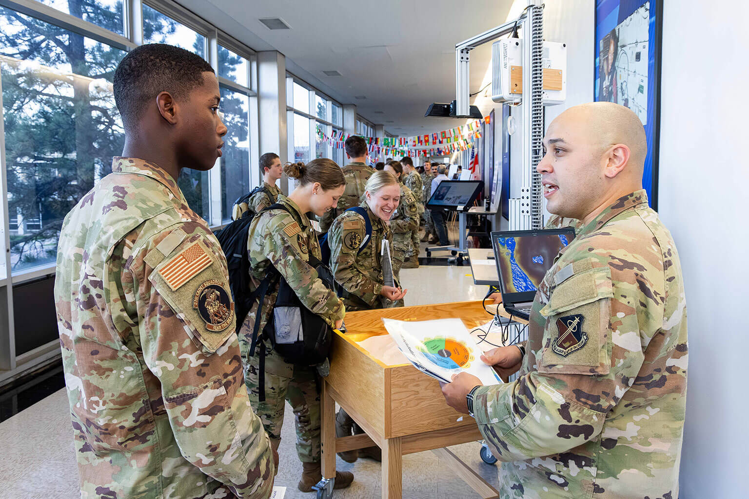 A cadet engages in lively conversation in a bright room with flags and tech displays. The atmosphere is upbeat and interactive.