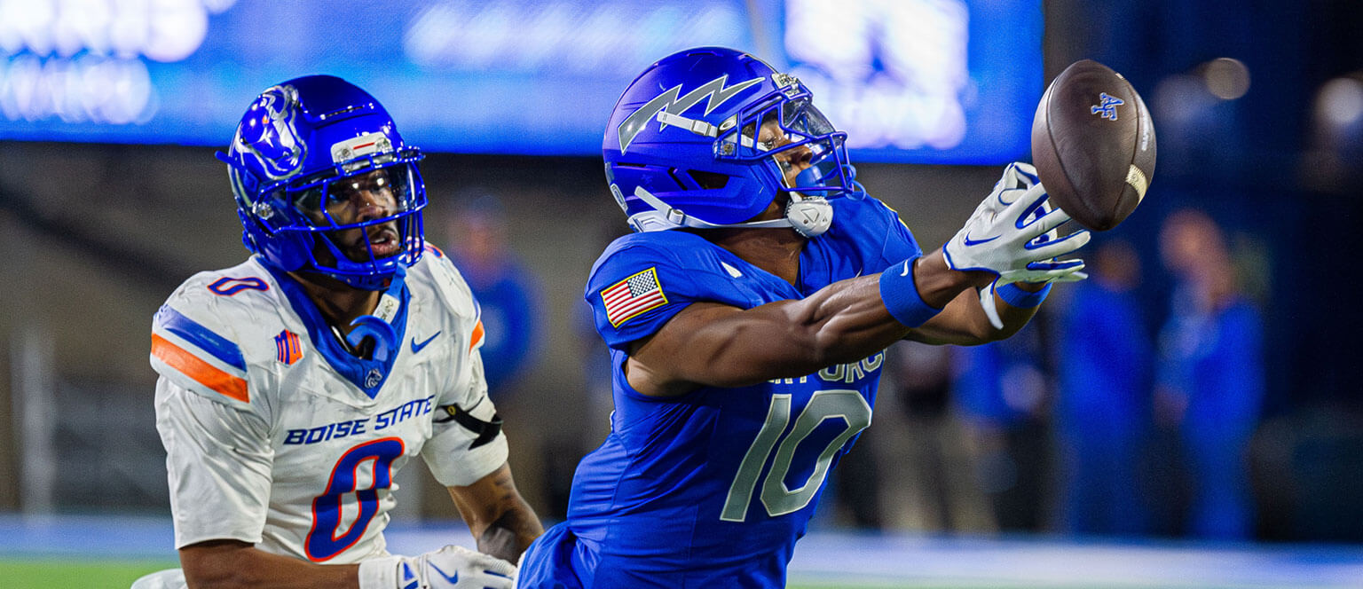 U.S. Air Force Academy player in blue successfully catches a football during a game with an opposing player in white uniform.