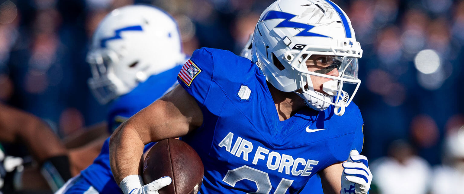 U.S. Air Force Academy football player in blue and white gear, actively running with a football during a game.