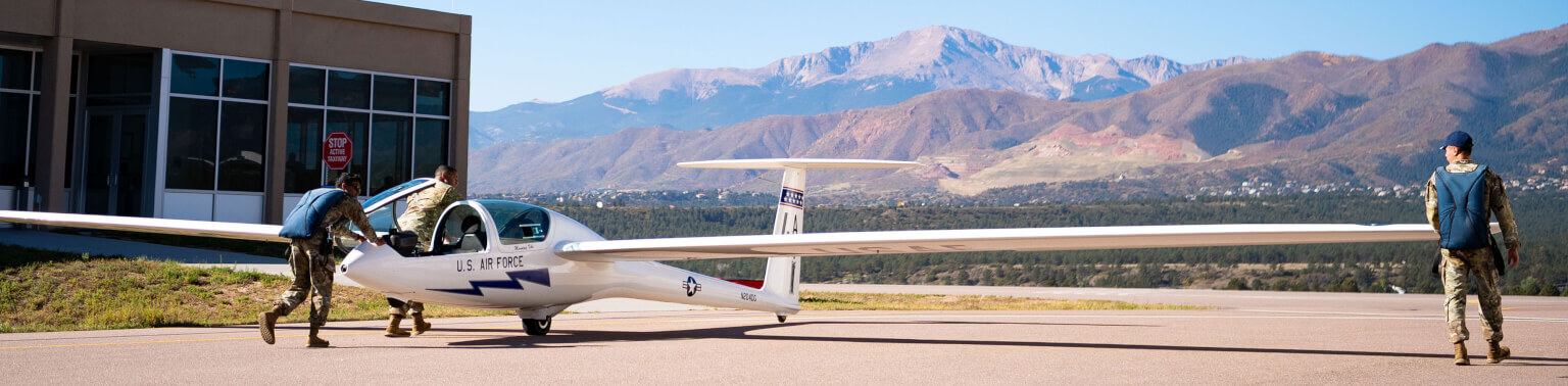 Three cadets in flight suits prepare a glider on an airport tarmac, with a mountainous landscape in the background under a clear blue sky.