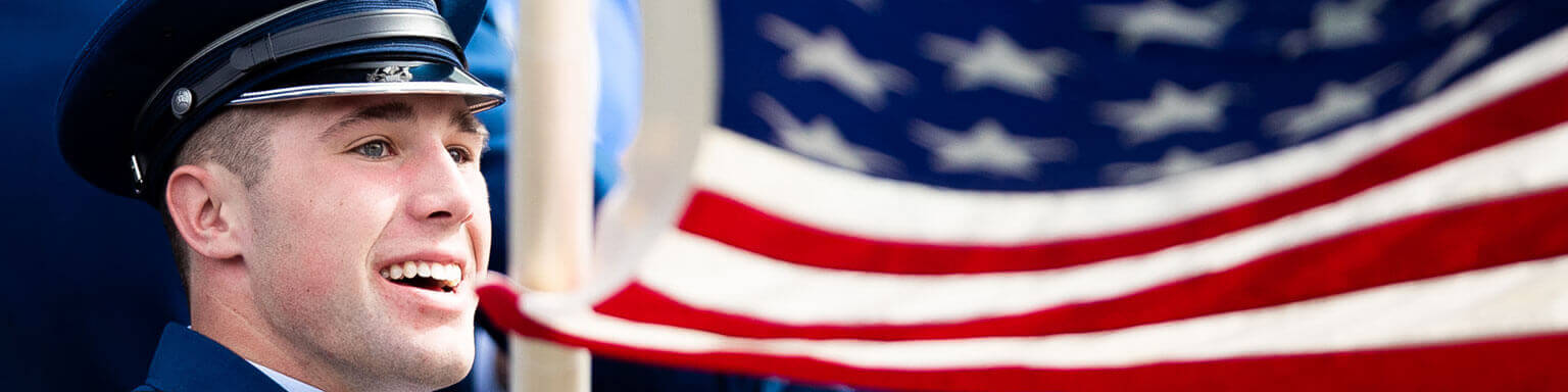 A smiling cadet in a blue military uniform stands beside a waving American flag, conveying patriotism and pride. The flag's stars and stripes are prominent.