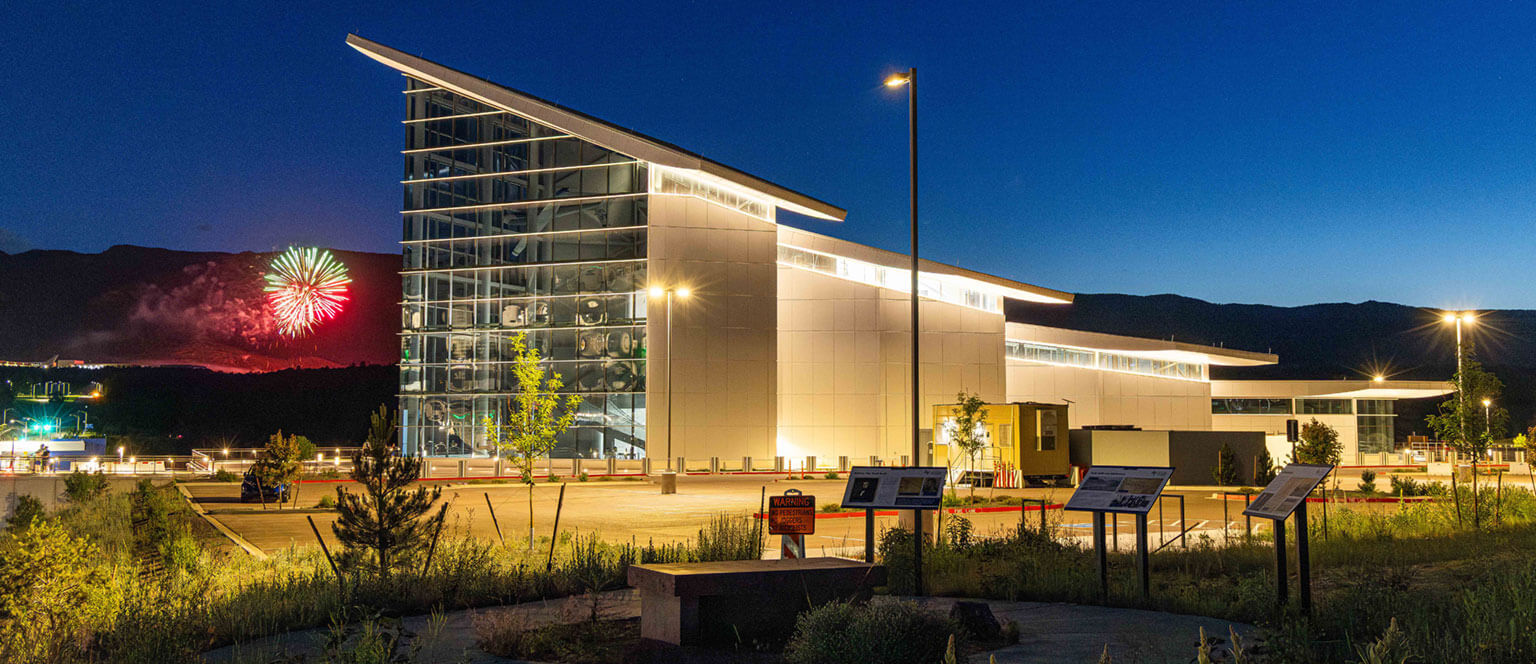The Hosmer Visitor Center is a modern building with large glass façade illuminated at night, set against a clear sky. Fireworks burst in the distance, adding a festive touch. Lush greenery and informational signs in the foreground enhance the scene.