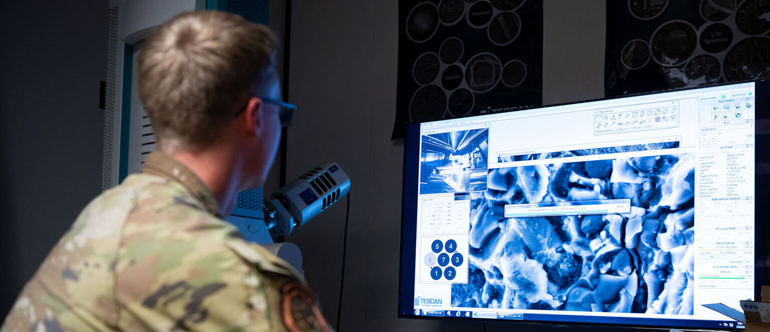 A cadet examines a computer displaying microscopic images in a lab. The setting is dim with a focus on scientific research and analysis.