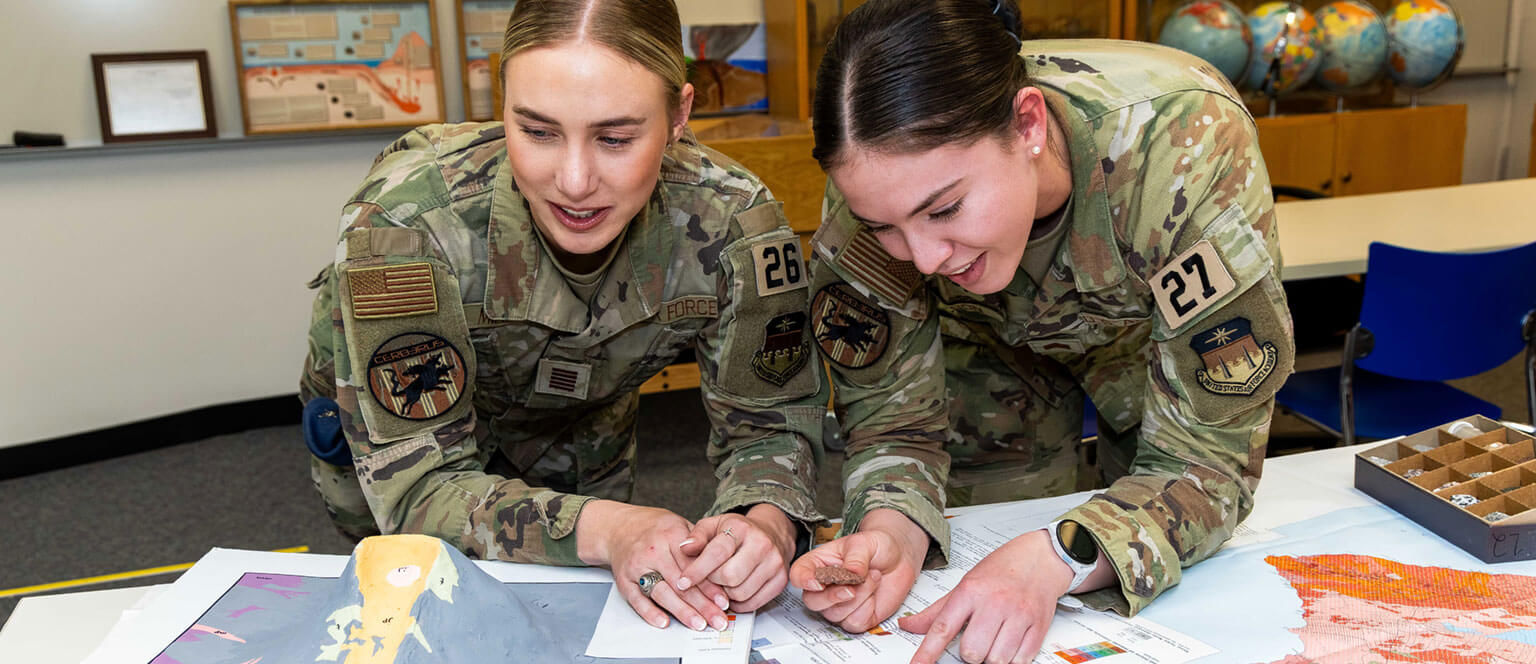 Two cadets in operational camouflage pattern uniforms lean over a table, examining maps enthusiastically. Globes are visible in the background, suggesting a classroom setting.