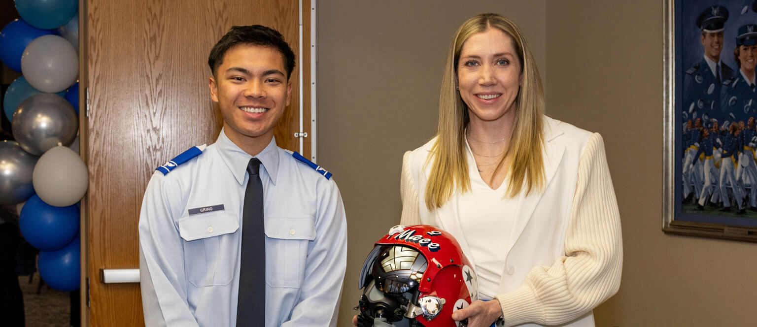 A U.S. Air Force cadet stands next to Maj Michelle “Mace” Curran holding a red helmet. Blue and white balloons and a military painting are in the background, conveying an official yet celebratory tone.
