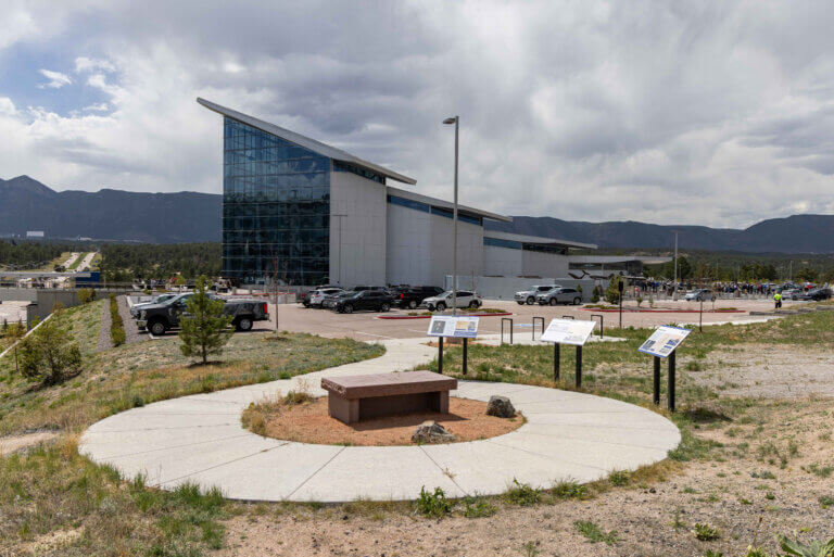 The Hosmer Visitor Center with large glass facade in front of mountains. Foreground features an outdoor display area with informational signs and a bench. Cloudy sky.