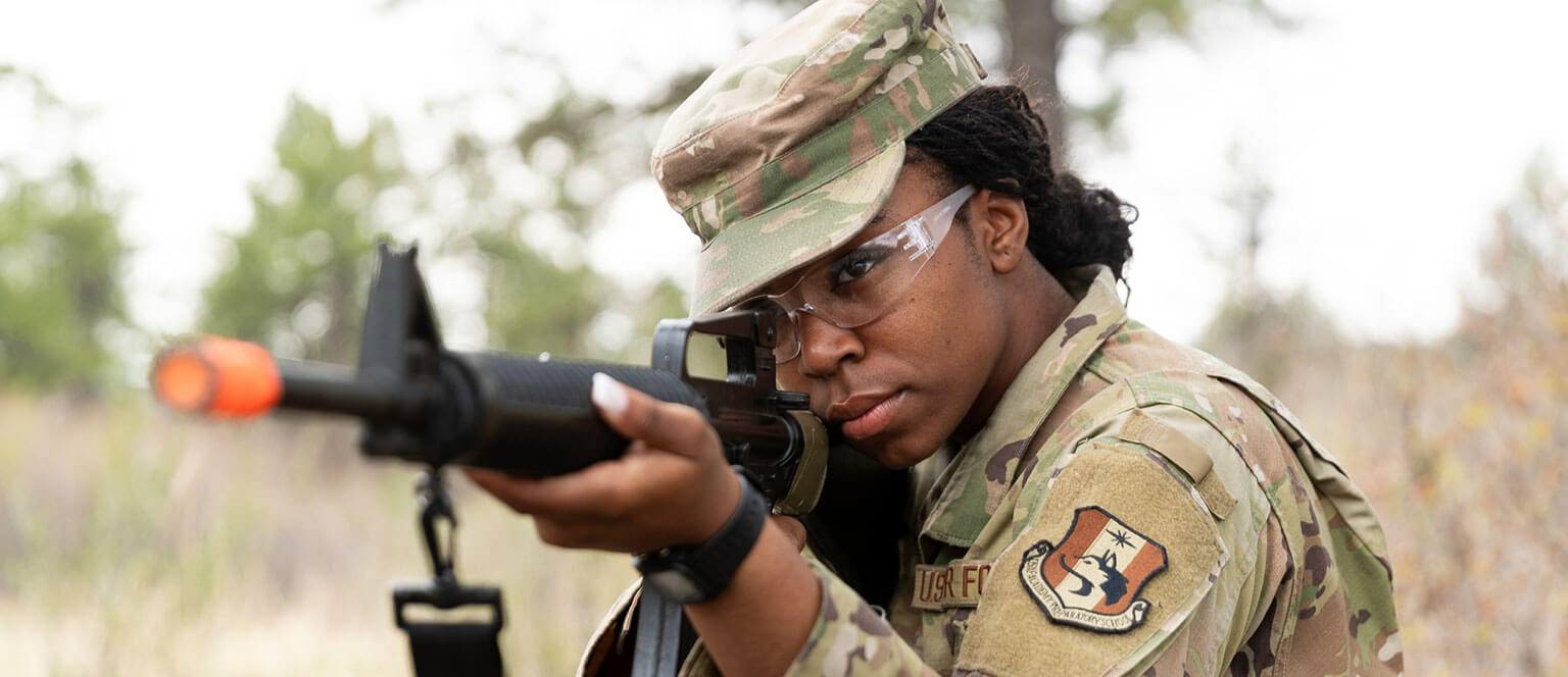 A U.S. Air Force Academy Preparatory School cadet candidate in military uniform and safety glasses aims a rifle outdoors, with trees and grass in the background.