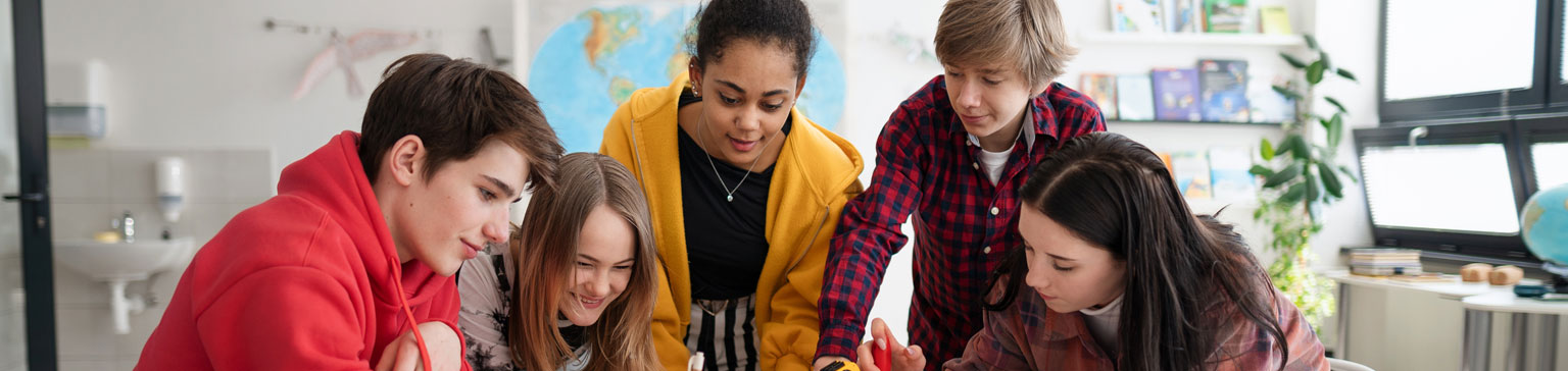 A diverse group of five engaged students collaborate at a desk, surrounded by books and a globe, conveying teamwork and learning in a classroom.
