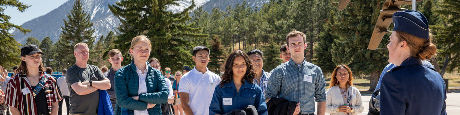 A group of young perspective candidates attentively listens to a speaker in uniform outdoors, with a mountainous backdrop and trees. The setting appears educational and engaging.