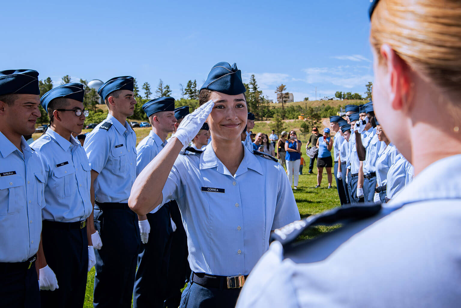 A group of uniformed cadets stands on a grassy field under a clear blue sky. One smiling cadet salutes another.