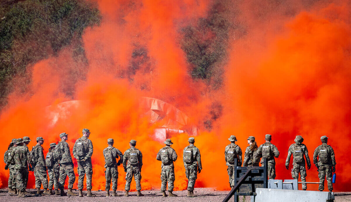 Cadets and cadre members stand near orange flare smoke during combat survival training