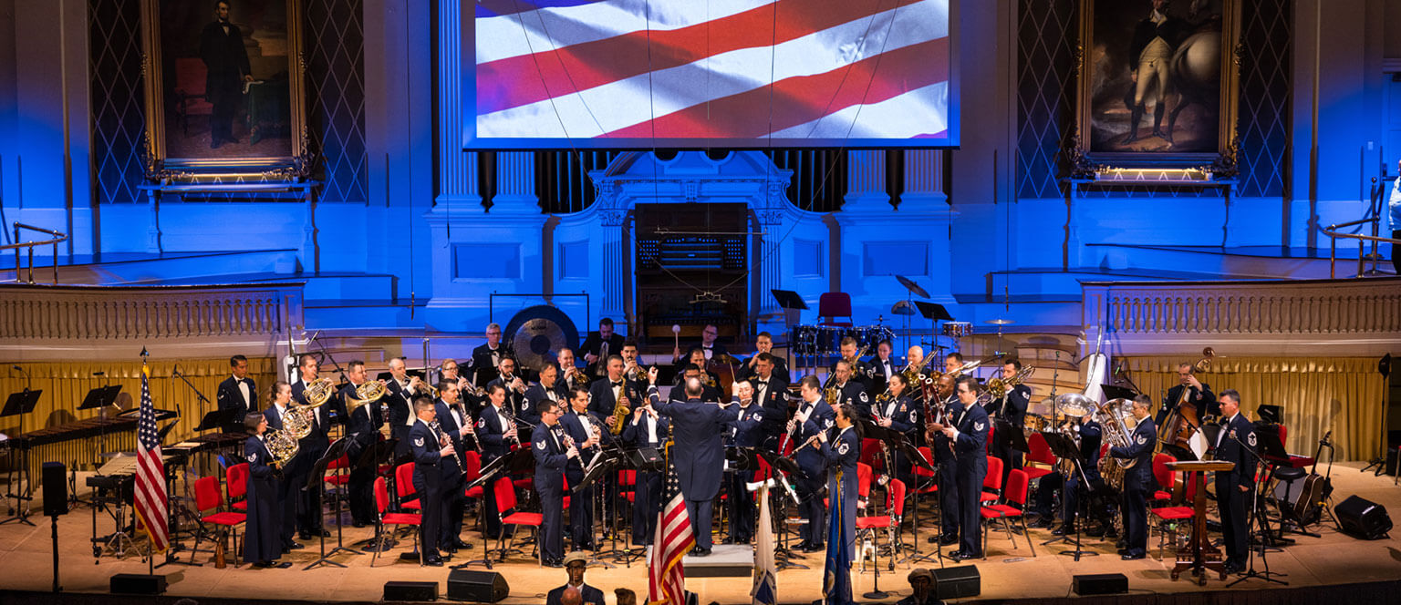The U.S. Air Force Academy band performs on stage, surrounded by U.S. flags. A large screen displays the American flag, creating a patriotic atmosphere.