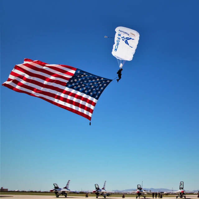 Wings of Blue member trailing US flag above Thunderbirds on airfield