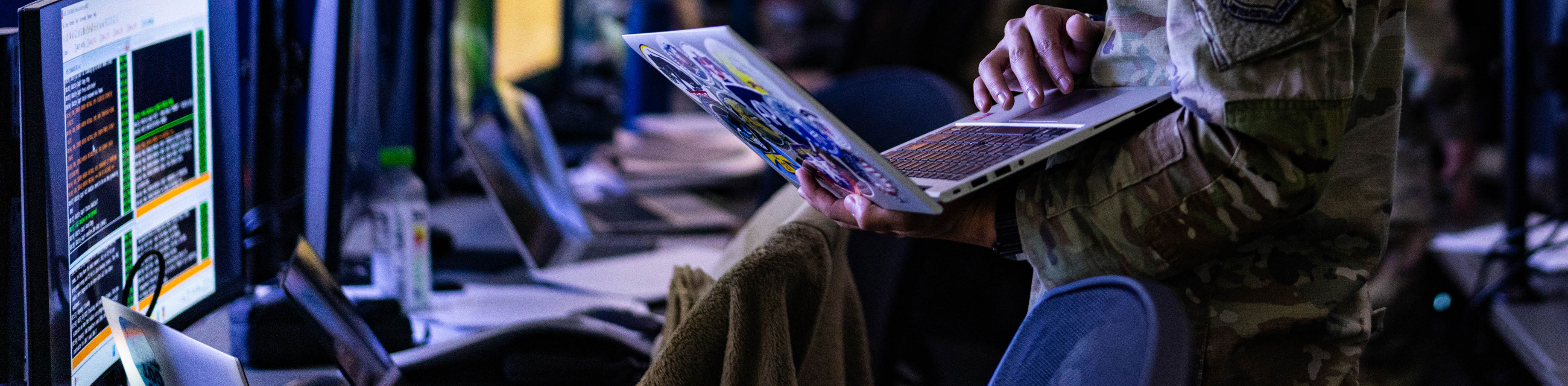 A cadet in military uniform works in a tech environment, using a laptop amidst a row of glowing monitors displaying code, conveying focus and technology.