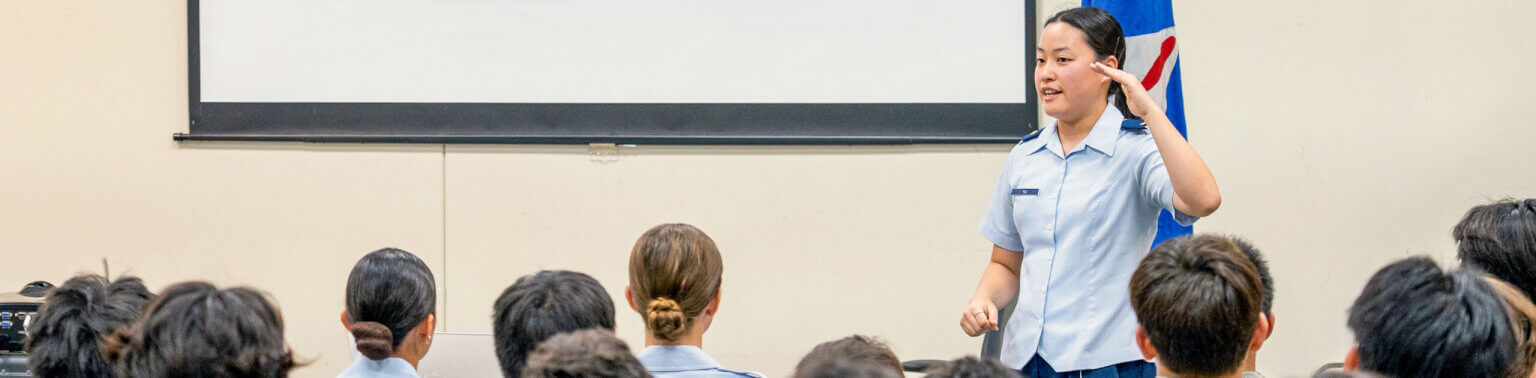 An officer in uniform speaks to a seated audience in a classroom setting, gesturing with her hand. A screen and flag are visible in the background.