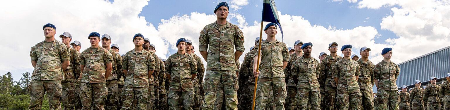 A group of cadets stand in formation outdoors, wearing camouflage and blue berets. The sky is partly cloudy, creating a formal and proud atmosphere.