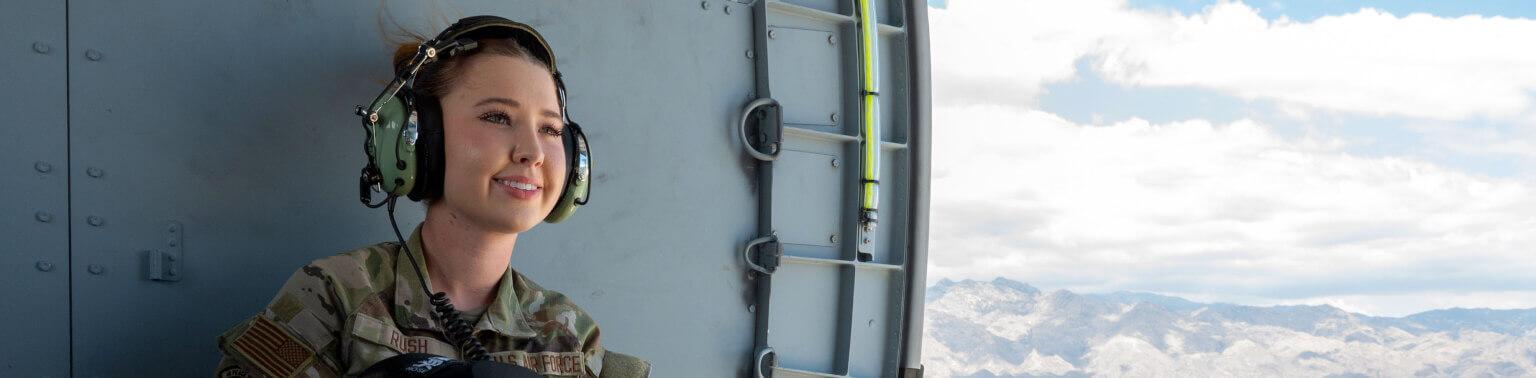 Young woman in military uniform and headset gazes out of helicopter door, looking content. Mountains and cloudy sky are visible in the background.