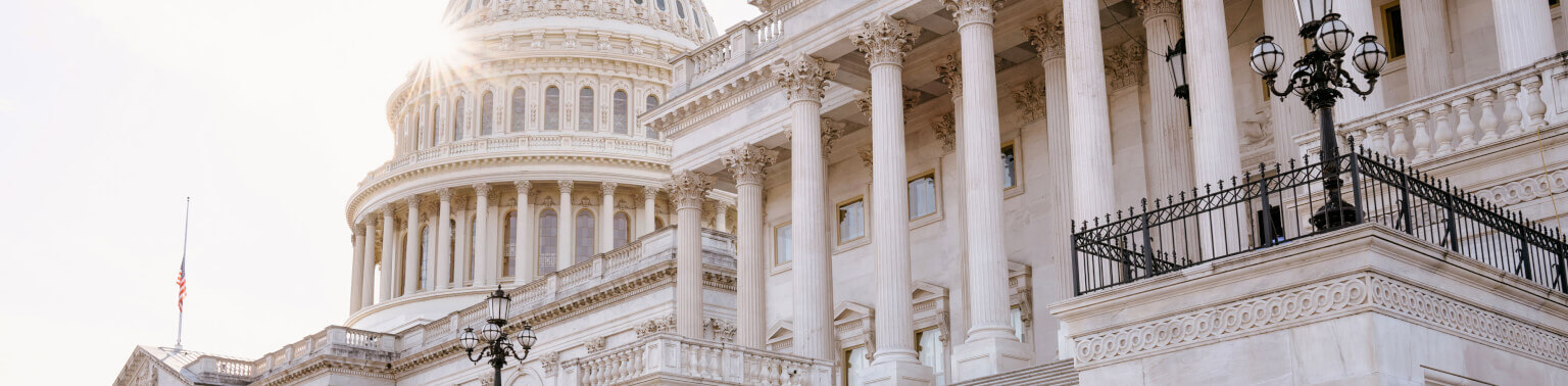 The image shows the U.S. Capitol building in bright sunlight. The dome is prominent, with classic white columns. An American flag is visible on the left.